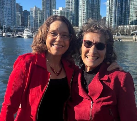Two women smiling by waterfront with cityscape background.