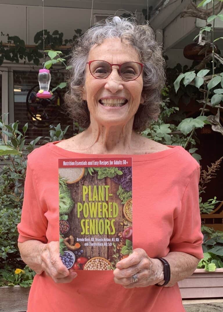 Smiling woman holds Plant-Powered Seniors cookbook in lush garden.